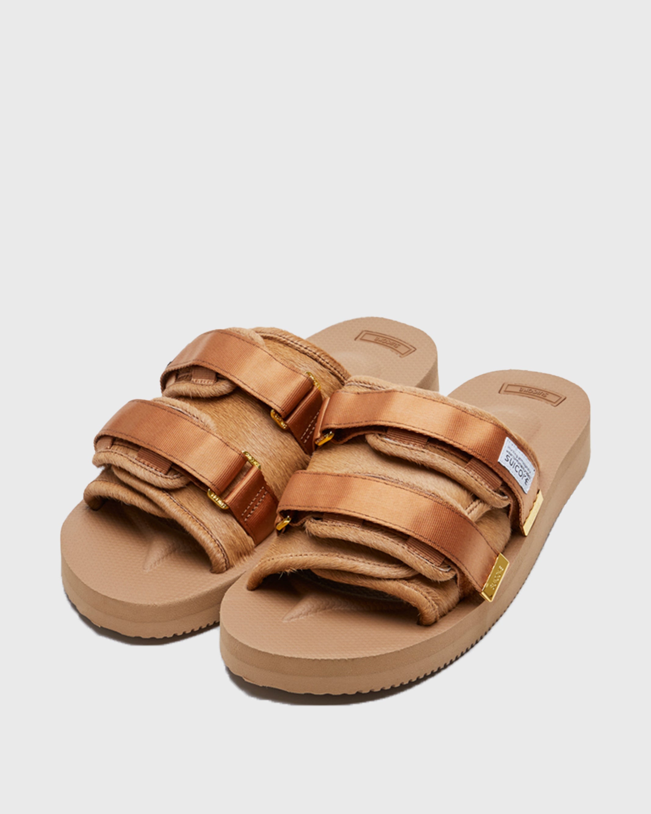 SUICOKE MOTO-Vhl slides with camel colored calf hair upper, camel colored midsole and sole, straps with logo patch and gold logoed tabs. From Fall/Winter 2021 collection on SUICOKE Official US & Canada Webstore.