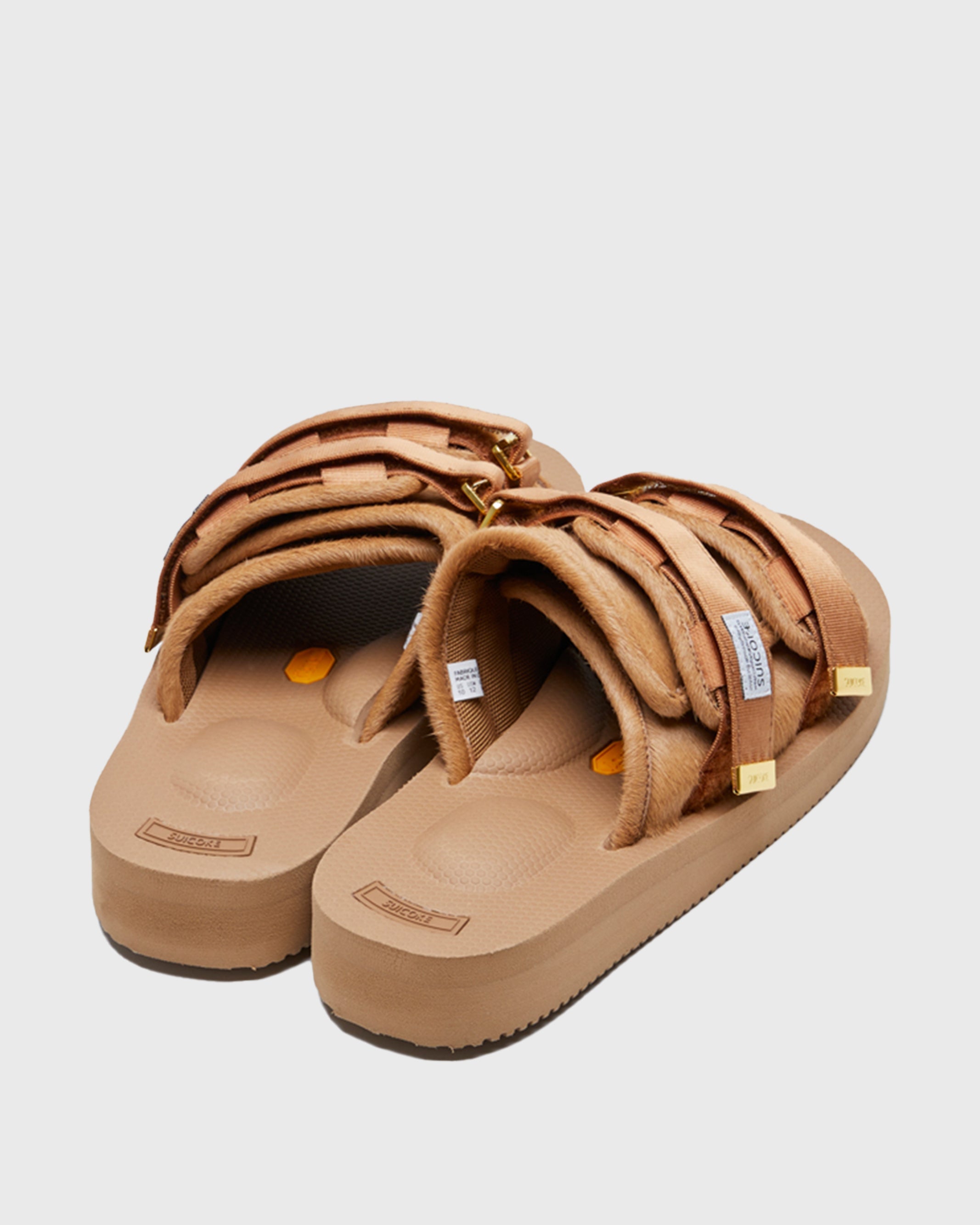 SUICOKE MOTO-Vhl slides with camel colored calf hair upper, camel colored midsole and sole, straps with logo patch and gold logoed tabs. From Fall/Winter 2021 collection on SUICOKE Official US & Canada Webstore.